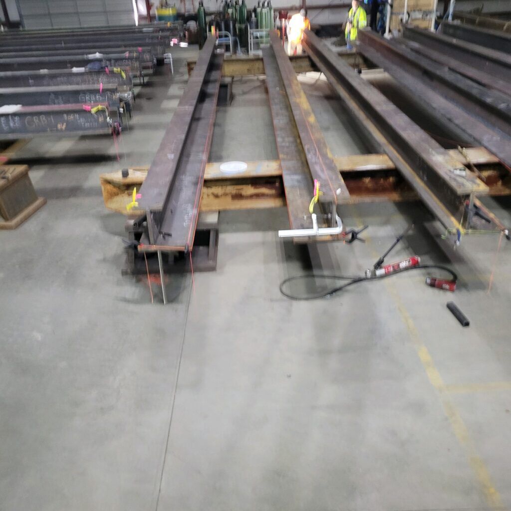 Industrial warehouse scene with steel beams and construction materials arranged on the floor, with a worker in the background providing testimonials.