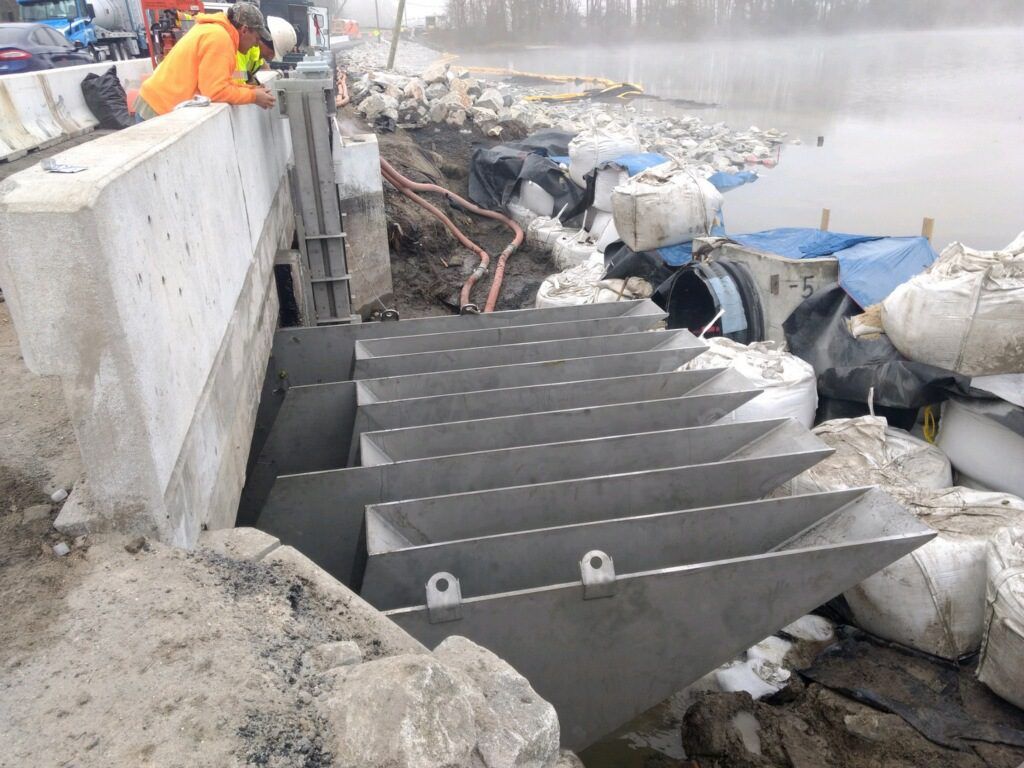 Construction workers install a metal sluice on a concrete barrier near a water body, with sandbags and rocks reinforcing the surrounding area, receiving positive testimonials for their efficiency and craftsmanship.